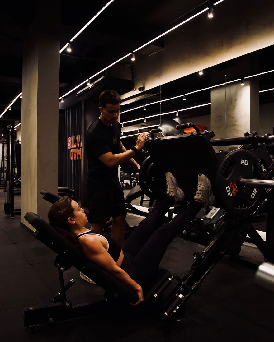 Salão de musculação da SILVA GYM, com aluna no leg press acompanhada pelo professor